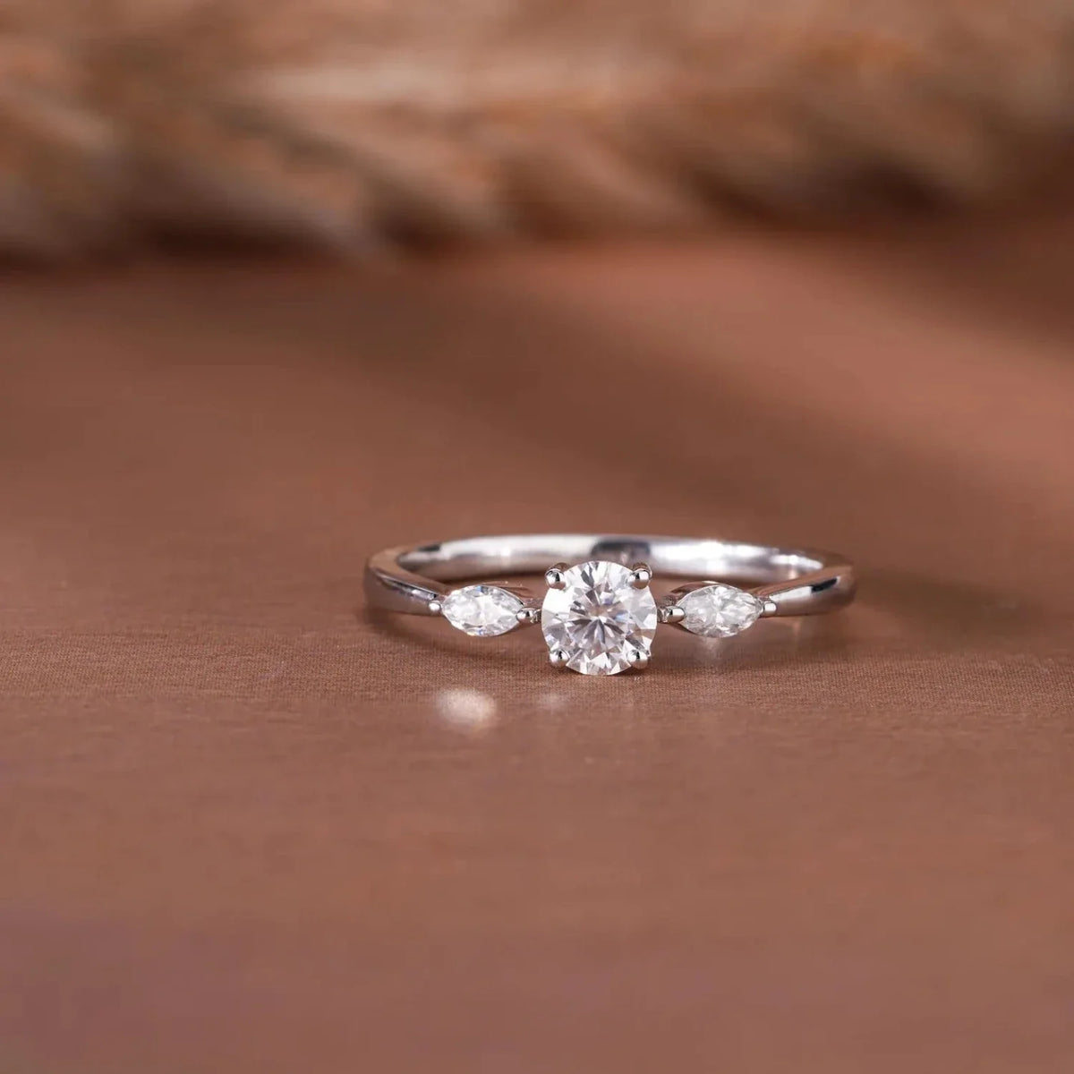 A silver three-stone ring with a round center gemstone and marquise-cut side stones, displayed on a warm brown surface with a soft blurred background.