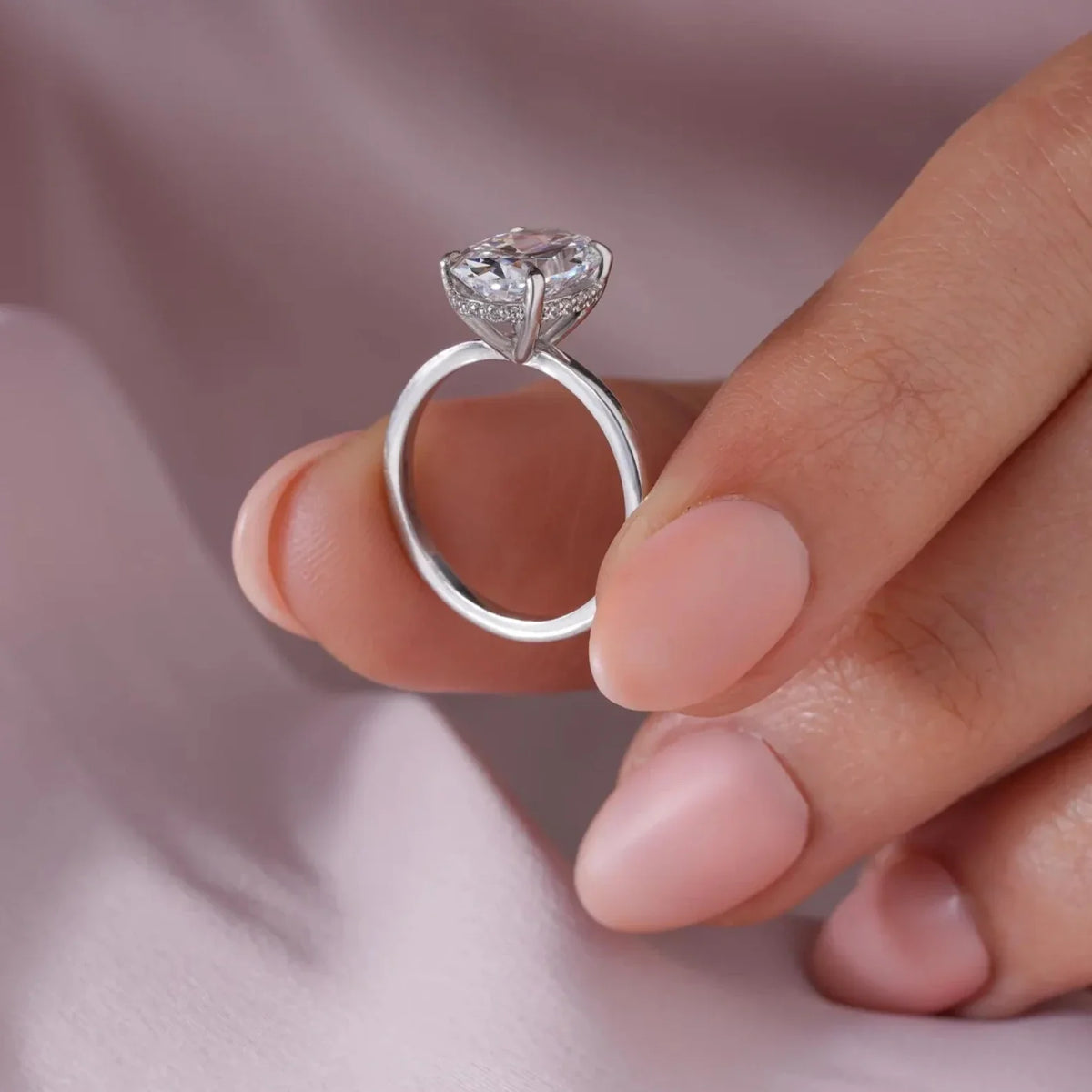 An oval-cut diamond ring on a silver band, shown worn on a person's hand resting on soft pink satin fabric. The brand name NIRA is visible in the bottom right corner.
