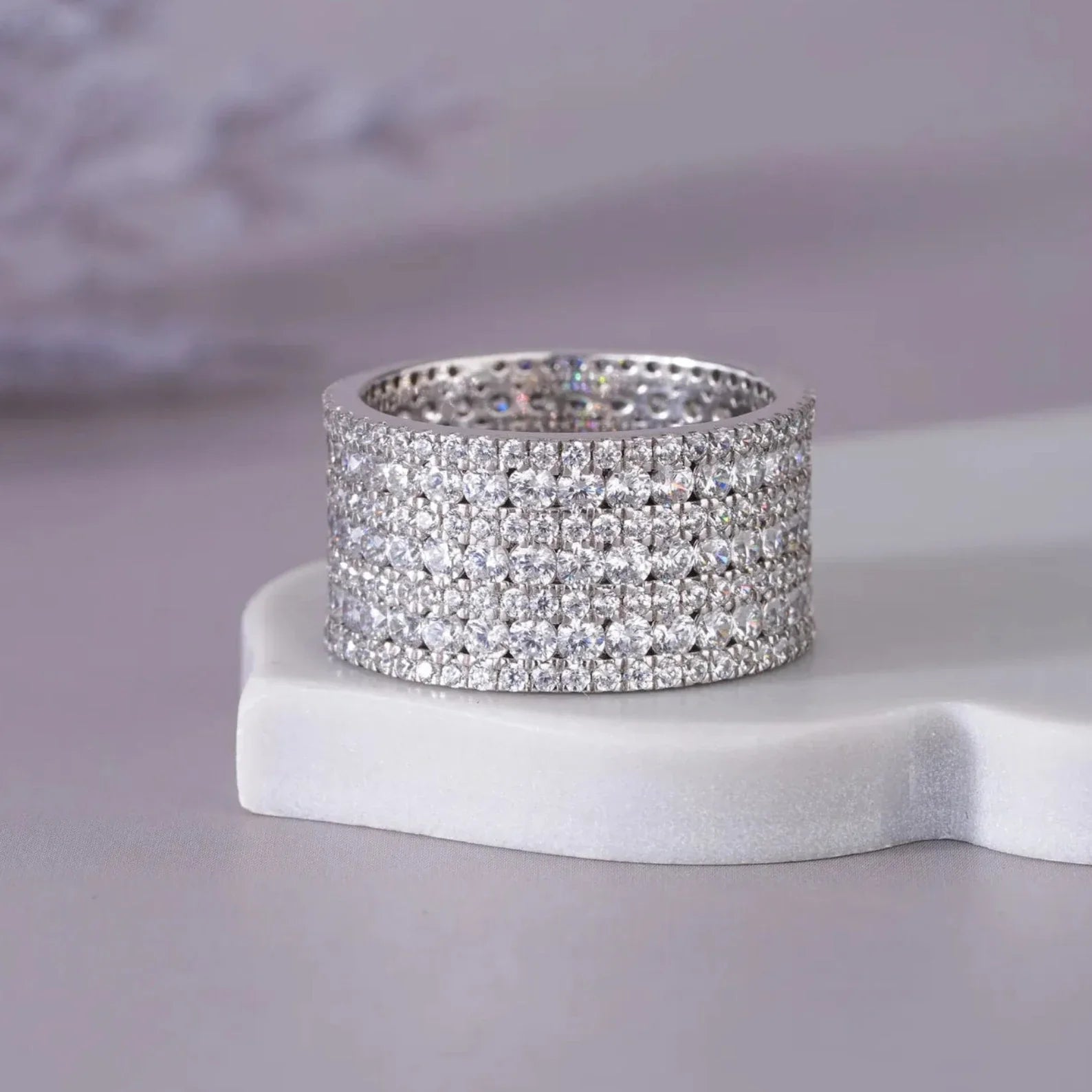 A wide silver-toned ring with three rows of sparkling clear stones, displayed on a white sculptural stand against a soft lavender-gray background. The brand name NIRA is visible in the bottom right corner.